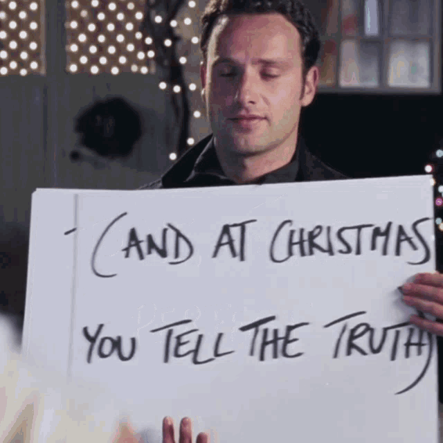 The image is from the movie Love Actually, featuring Andrew Lincoln's character, Mark, holding up a large sign that reads, "And at Christmas, you tell the truth." This scene is part of Mark's silent confession of love to Keira Knightley's character, Juliet.