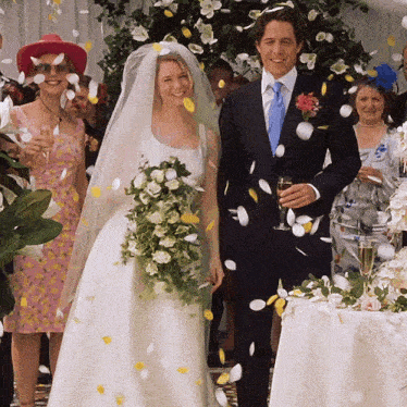 The image shows a wedding scene from Bridget Jones's Diary featuring Bridget Jones, played by Renée Zellweger, and Mark Darcy, played by Colin Firth. The couple is standing together, smiling as flower petals are tossed around them. Bridget is wearing a classic white wedding dress with a veil, and Mark is in a suit. Guests are celebrating in the background, adding to the joyful and festive atmosphere of the moment.