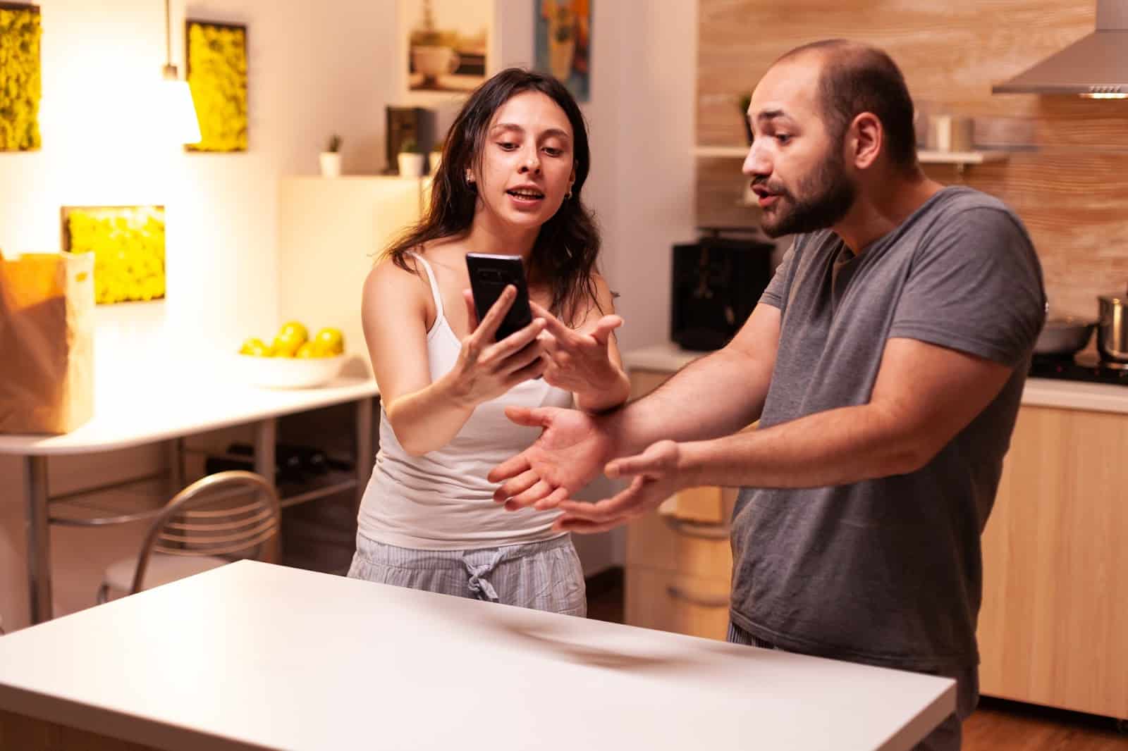 A couple is standing in a kitchen, engaged in a tense discussion. The woman is showing something on her phone to the man, who gestures as if explaining or defending himself. Both appear frustrated, suggesting a disagreement.