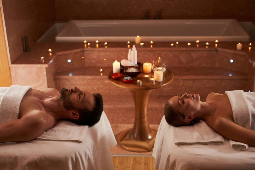 The image shows a couple lying on separate massage tables in a serene spa setting. Both are wrapped in white towels and appear relaxed, with their eyes closed. The ambiance is peaceful, with soft lighting and candles surrounding a wooden table between them. Various spa products and decorative candles are arranged on the table, enhancing the calming and luxurious atmosphere. A large bathtub is visible in the background, suggesting the spa experience includes multiple relaxation options.