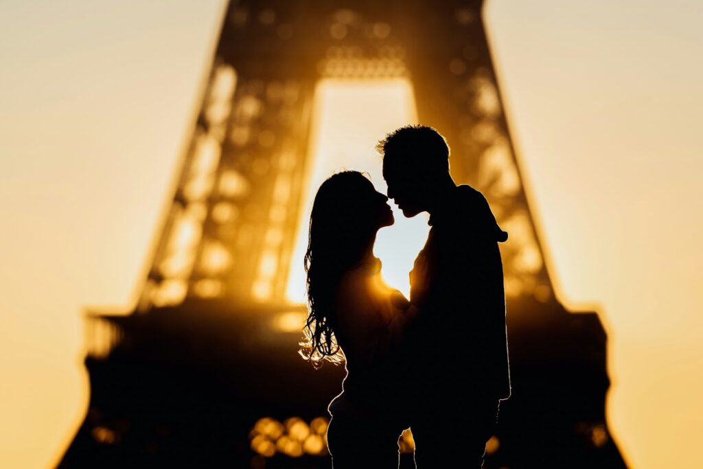 The image shows a romantic silhouette of a couple standing in front of the Eiffel Tower at sunset. The pair are about to kiss, with the warm golden light of the sunset creating a soft glow around them. The Eiffel Tower in the background adds to the iconic and romantic ambiance of the scene.