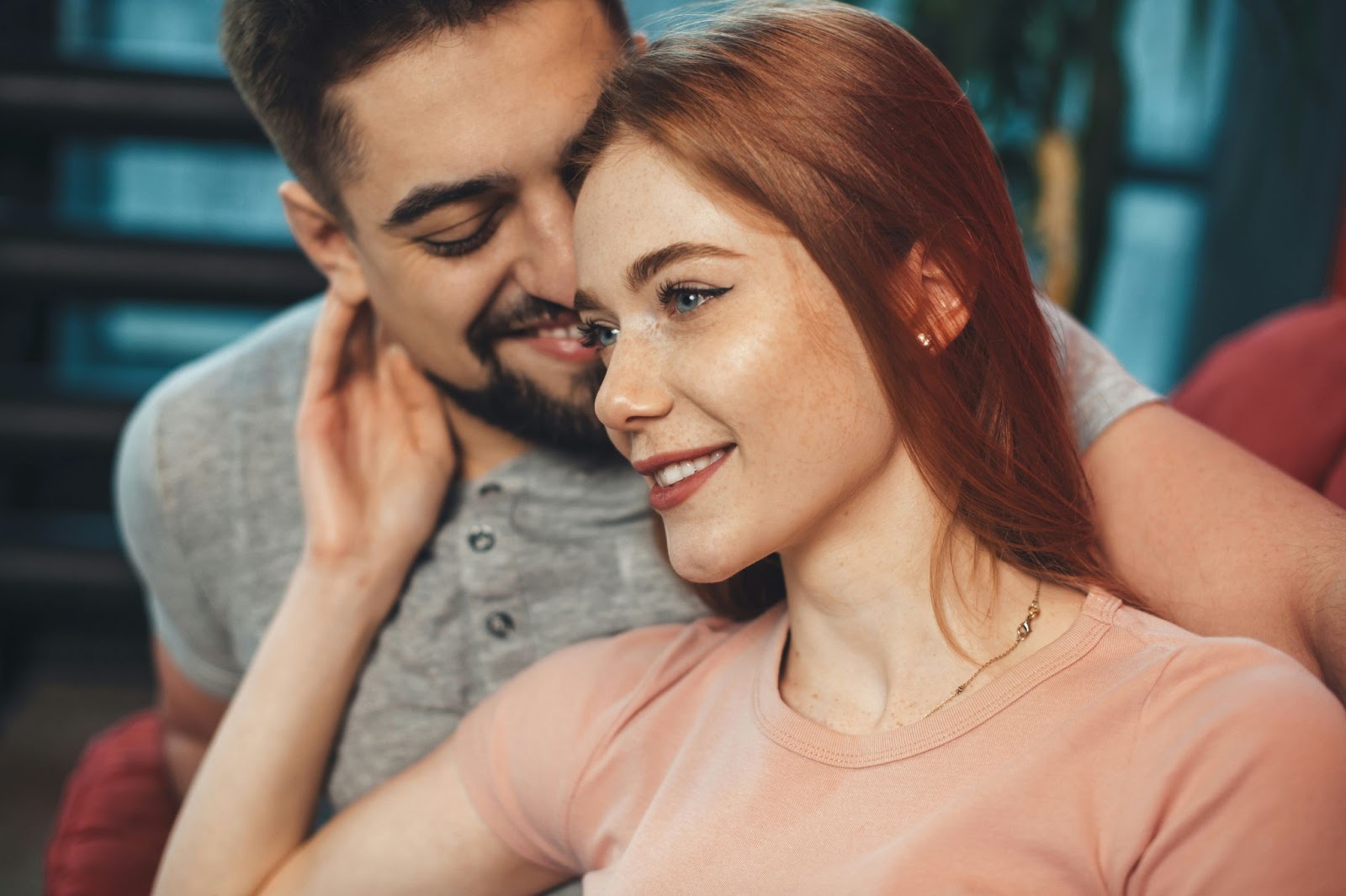 A young couple sitting closely together, with the man smiling affectionately toward the woman, who is gazing ahead with a soft smile. The woman has long red hair, and the atmosphere is warm and intimate.